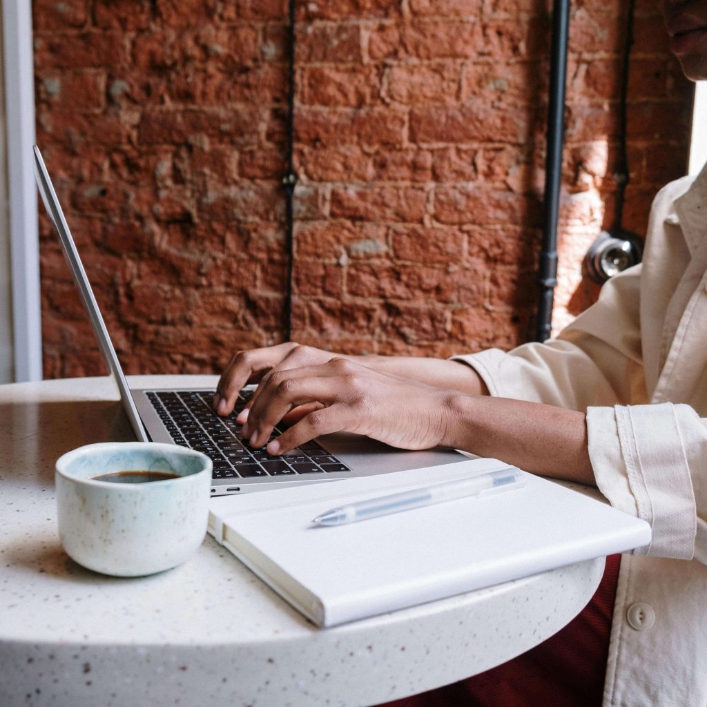 a women in a white shirt is sitting at the table at a cafe with her computer at the table and typing
