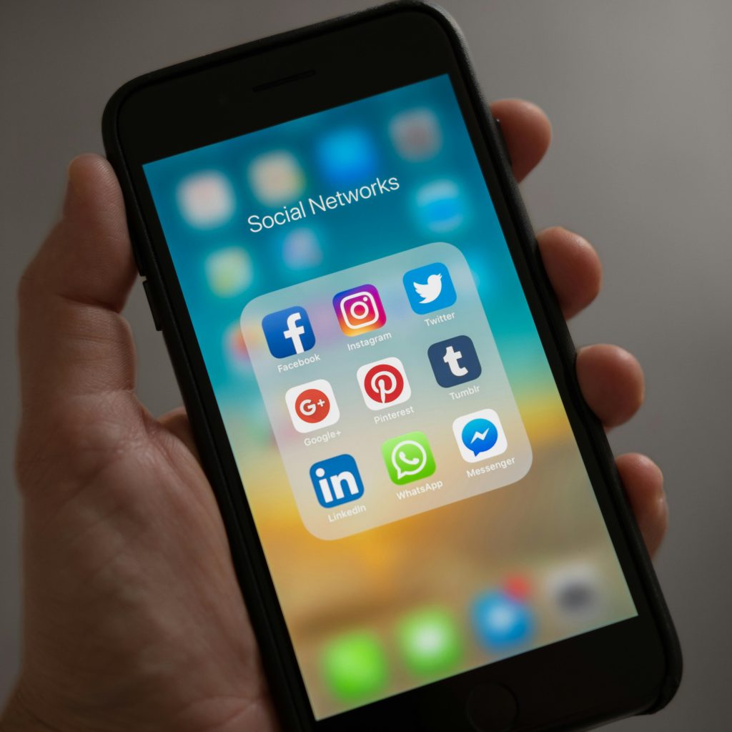 iphone in someones hand, with a folder of "social networks", including facebook, Instagram, twitter, google, pinterest, tumbrl, linkedin, whatsapp, messenger